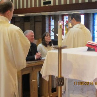People at a ceremony: couple, two clergy members in church. Candle lit on altar.