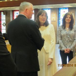 Two women and a man in a church, the man in a suit, the woman in a white dress, the other in a gray blouse and black pants.