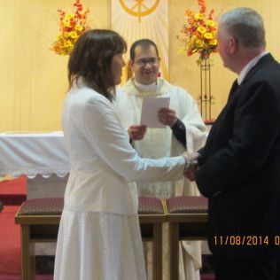 Couple exchanging vows in church with officiant; sunlight, flowers.