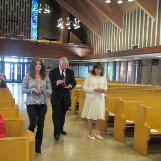 Three people walking inside a church, holding objects. Woman in white dress, man in black suit, another woman. Rows of pews.