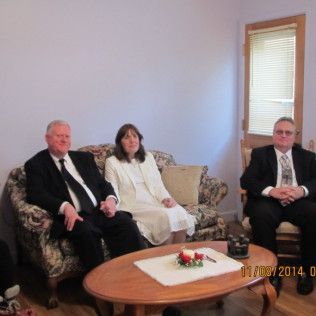 Three people in formal attire seated in a living room. Two on a floral couch, one in a chair.