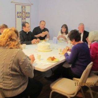 People seated around a table, eating and talking. A cake sits in the center. A cross is hanging on the wall.