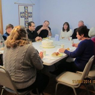 People seated around a table, possibly at a church gathering. There's a cake, food, and religious banner.