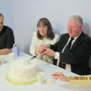 Man in suit cutting a cake with a knife, with two people at a table, and a small gift box.