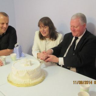 Three people cutting a cake at a table; the man on the right wears a suit, and they are smiling.