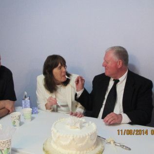 People at a table, eating cake; man in suit feeds woman, cake on table.