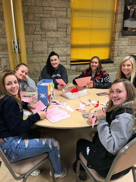 Six young people seated around a table, crafting. They are smiling and working with paper and supplies in a well-lit room.