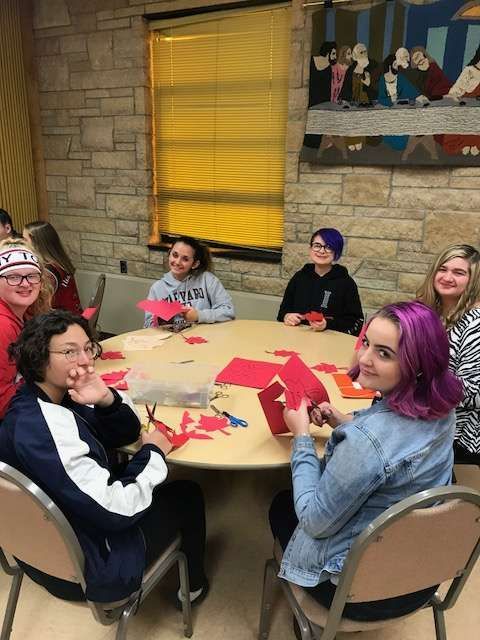Group of students at a table, crafting with red paper. Smiling faces, scissors, and a tapestry are visible.