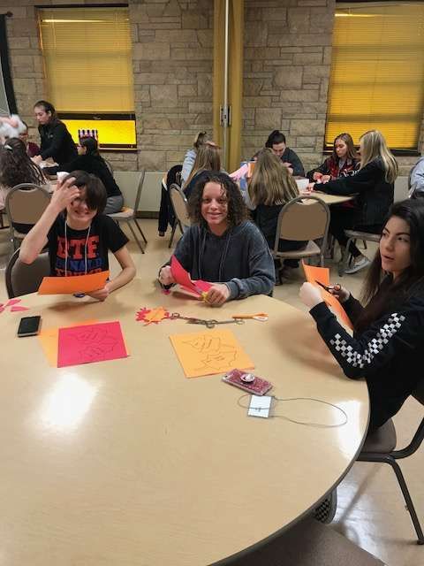 Students at a table craft with orange and red paper. One boy smiles, another covers his eyes. Others work in the background.