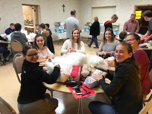 People seated at round tables sewing stuffed animals. Several others in the background. Room lit with overhead lighting.