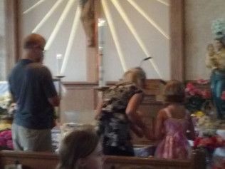 Family prays in a church. A woman and child kneel at an altar. Man stands beside them.