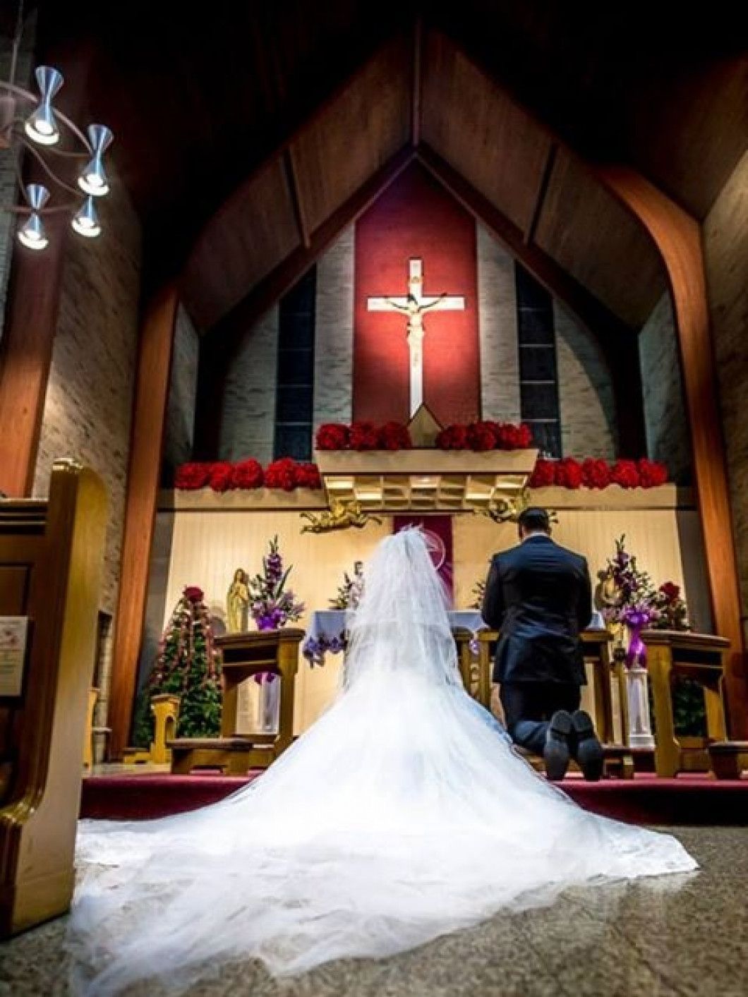 Couple kneeling at the altar in a church, the bride in a long white dress, a cross overhead.
