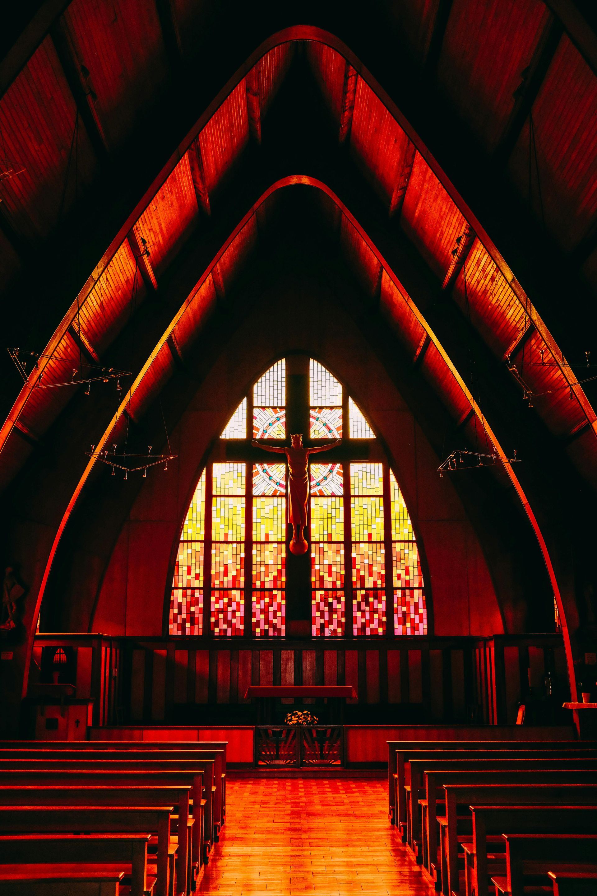 Interior church view with arched ceiling, stained-glass window, and wooden pews. Warm red and orange tones.