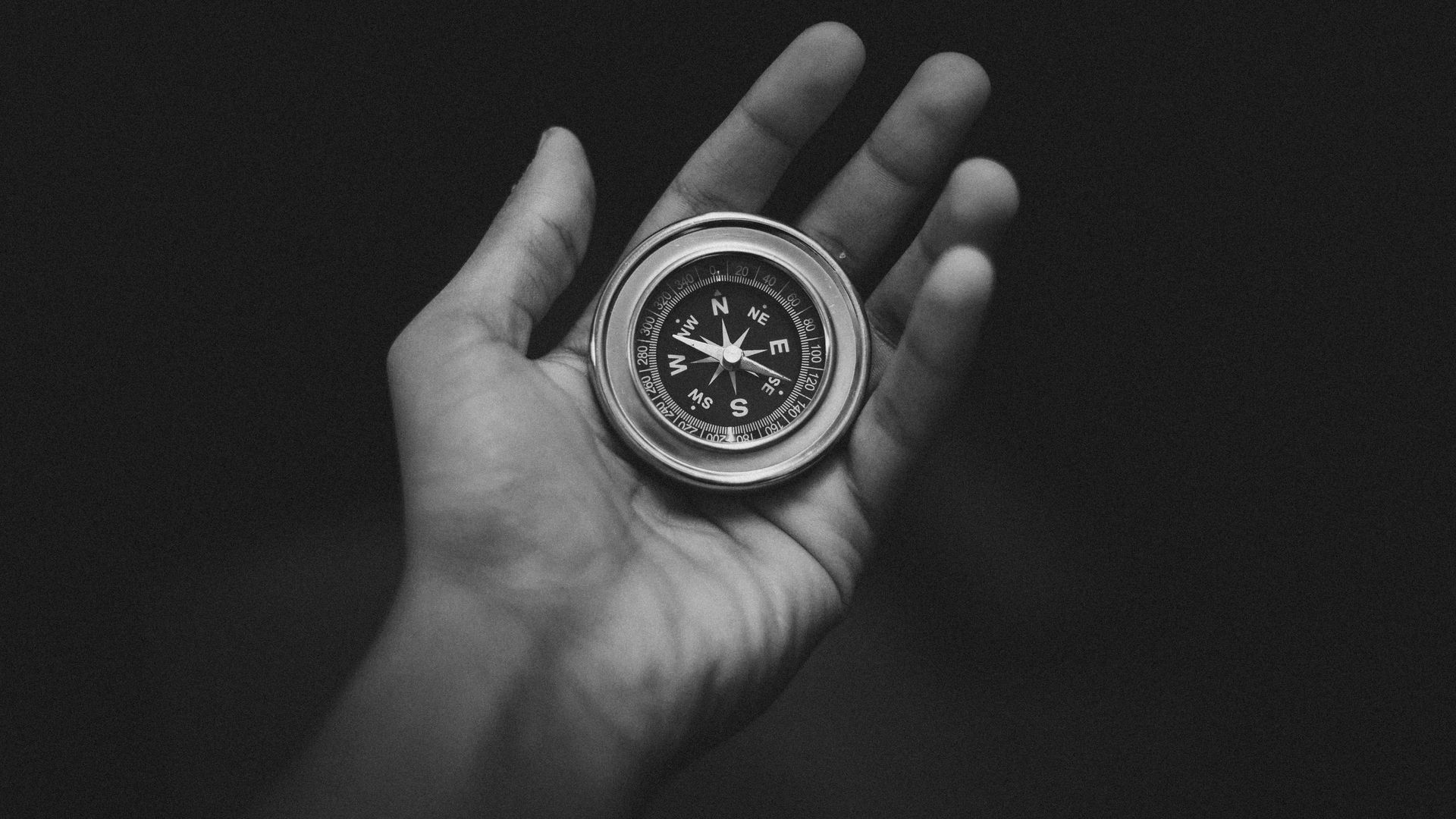 Hand holding a compass; the needle points north. Black and white.
