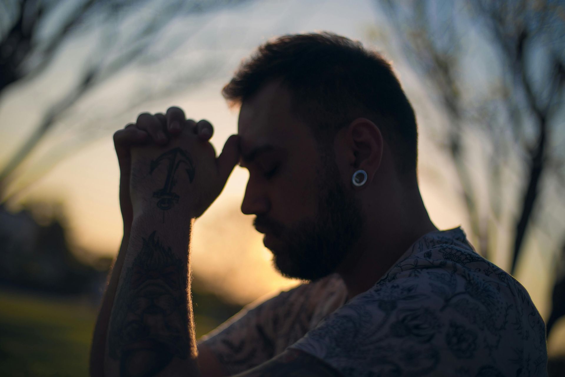 Man with tattoos, eyes closed, hands clasped, forehead on hands, sunset in background.