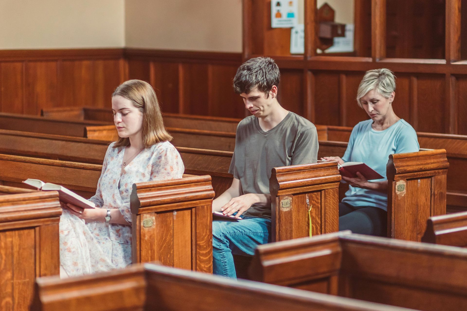 Three people reading books in wooden pews inside a church, lit by natural light.