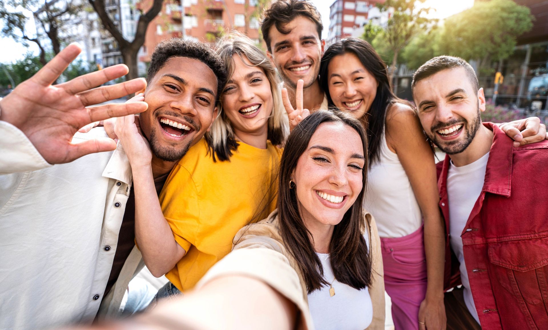 Group of smiling people taking a selfie outdoors; some are waving.