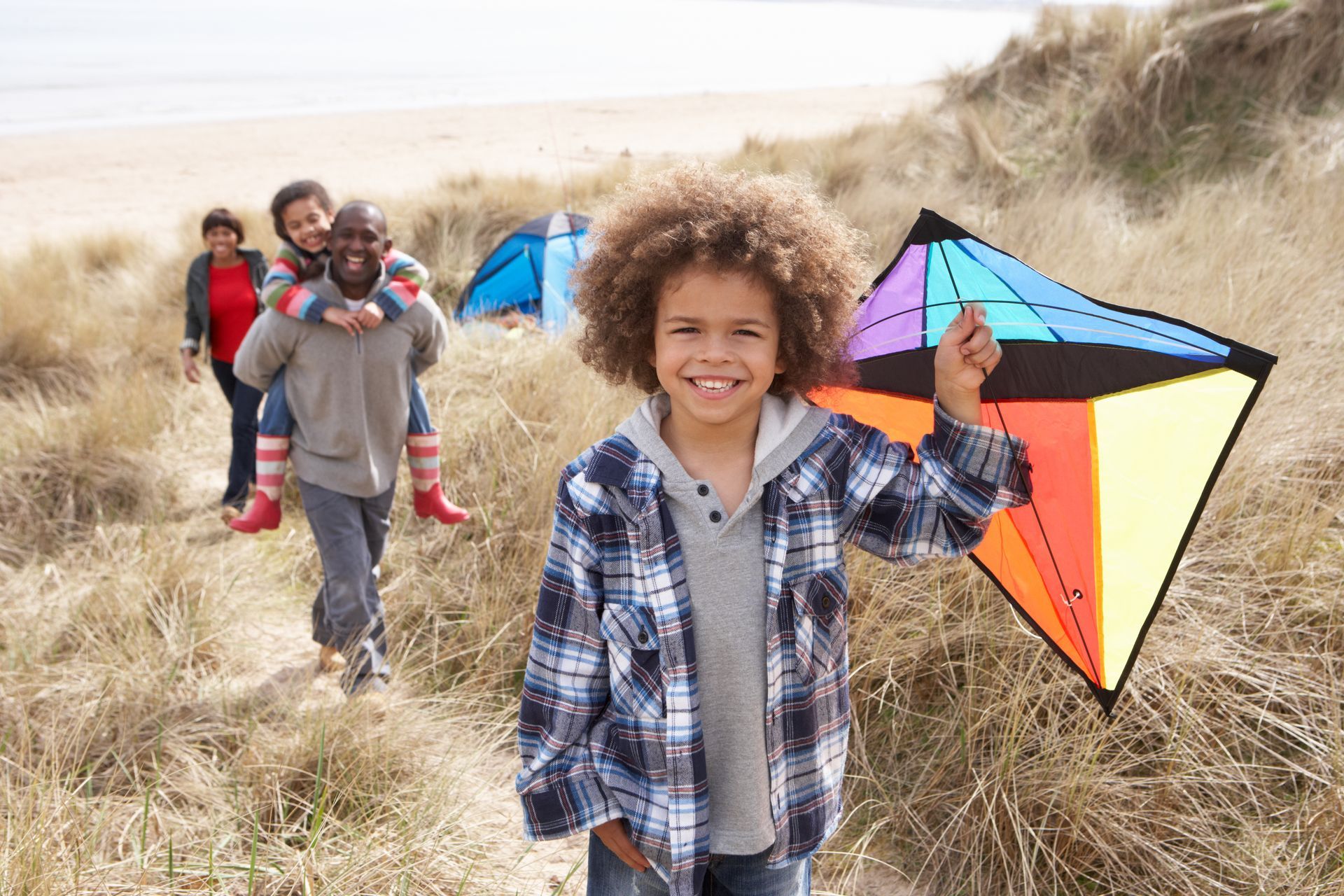 Boy holding a colorful kite, smiling at the camera, with family and tents in the background at a beach.