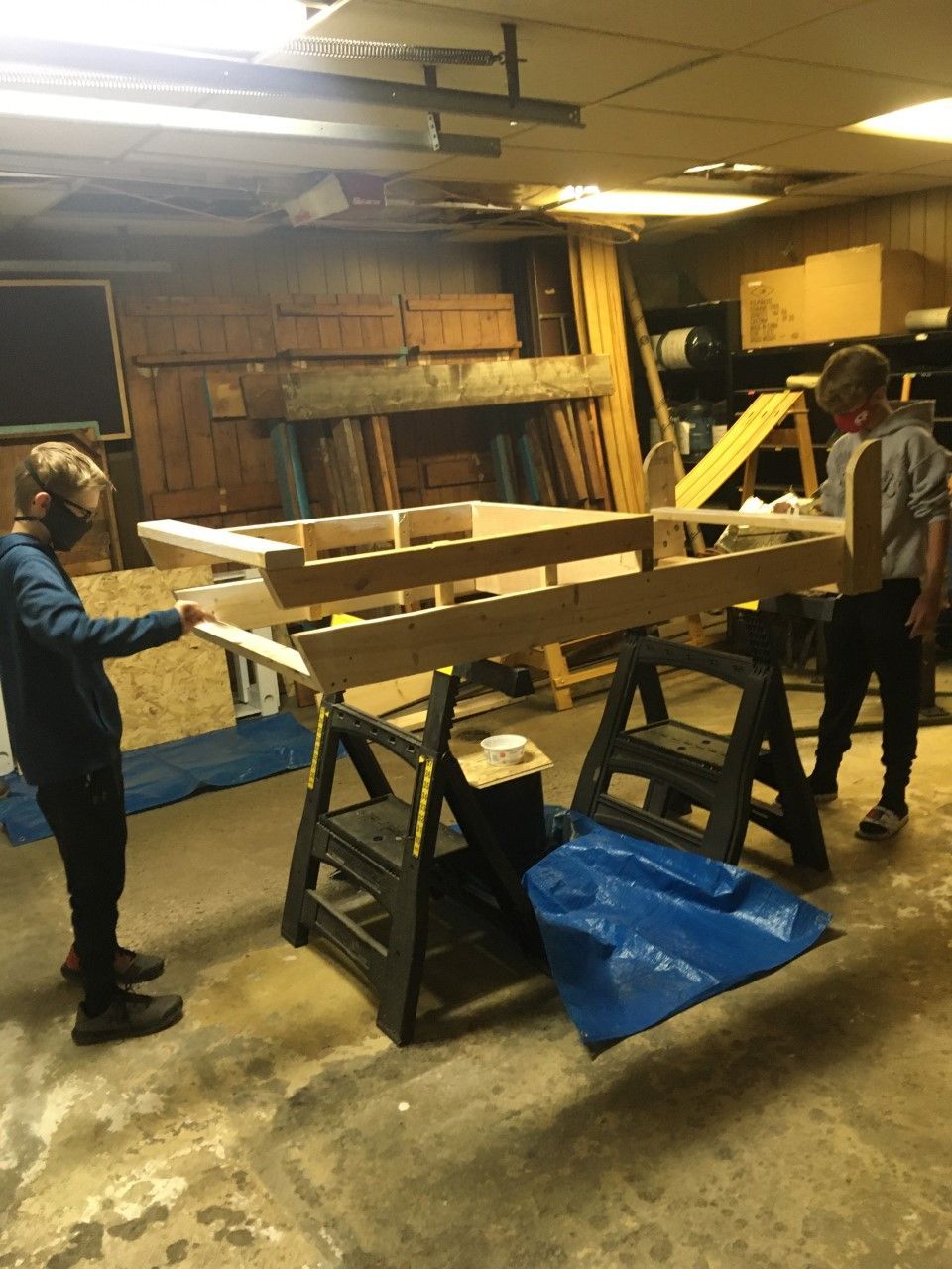 Two people building a wooden structure in a cluttered workshop, using sawhorses.