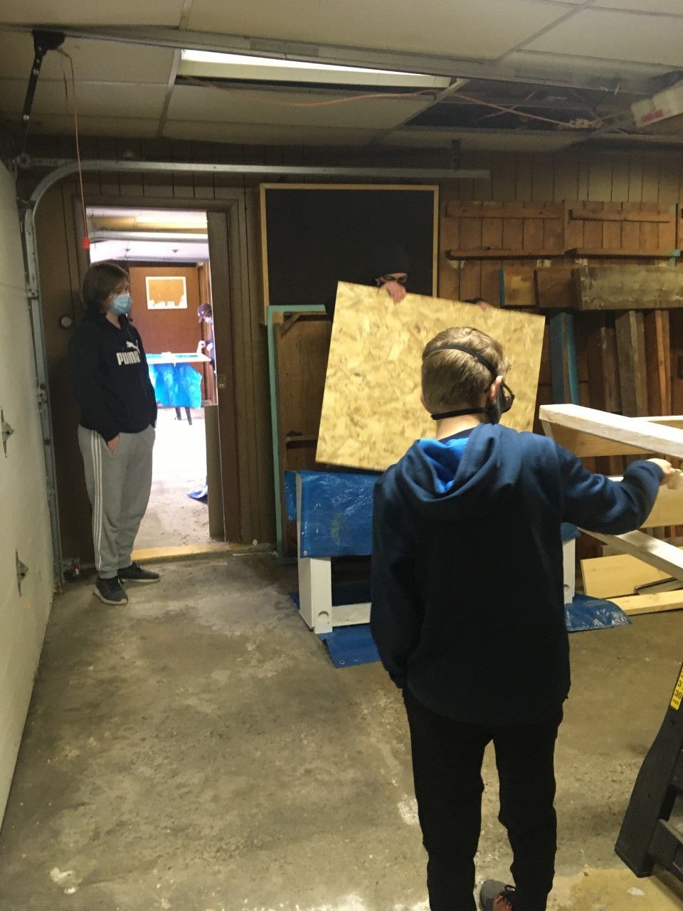 Two people in a workshop, one wearing ear protection, holding wood. Another stands in doorway wearing a mask.