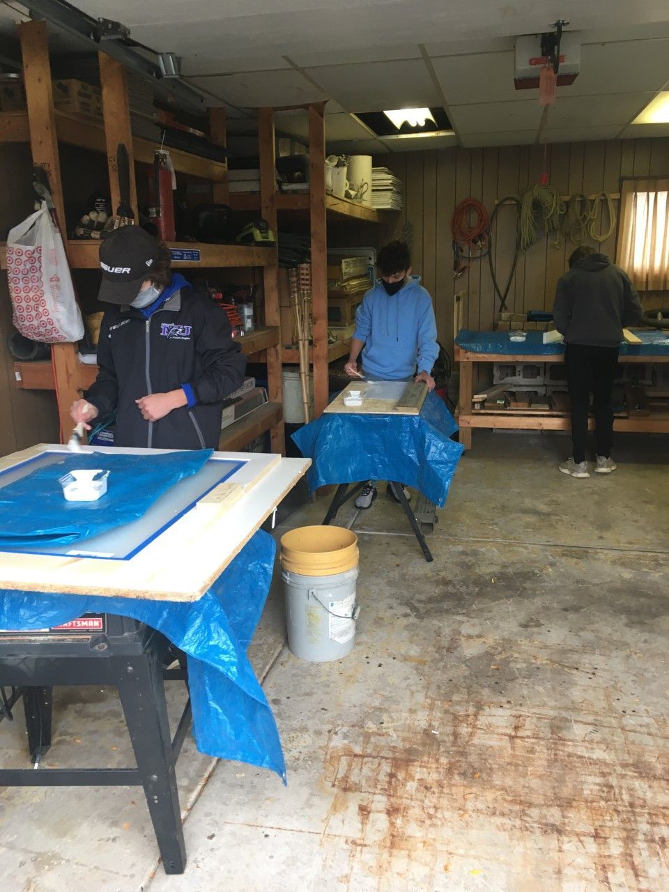 Three people painting inside a workshop. They wear masks and work at tables. Blue tarps cover surfaces.