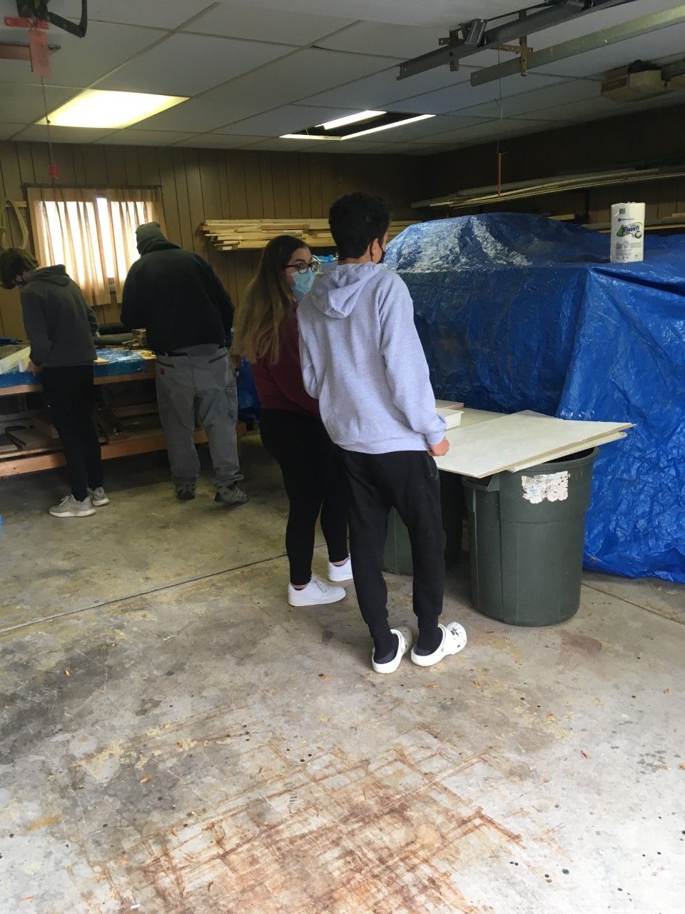 People working on a woodworking project in a workshop. A blue tarp and trash can are in the background.