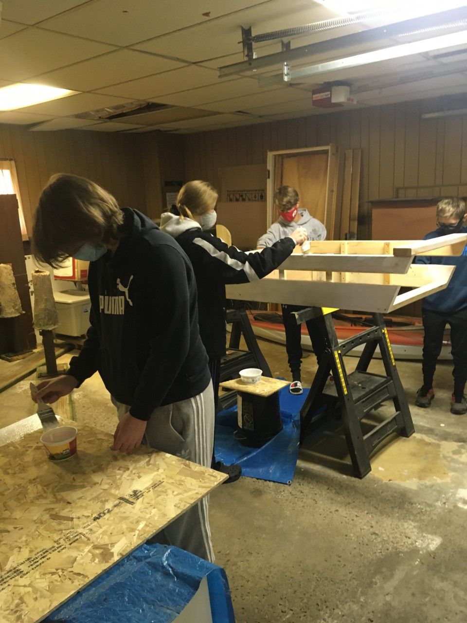 People painting a wooden structure in a workshop setting, wearing masks, using brushes.