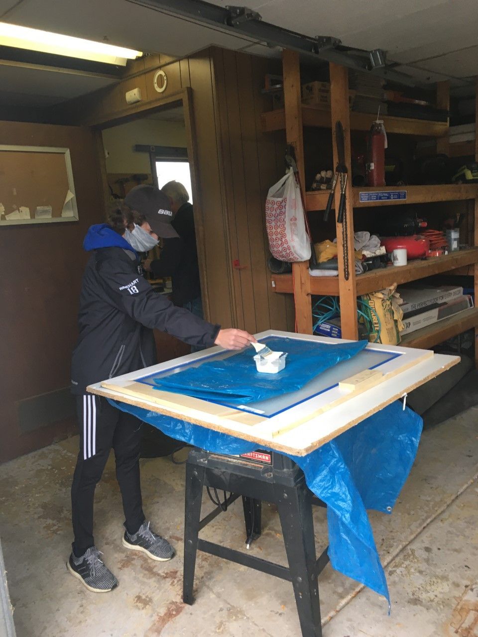 Person in mask works on a blue surface on a table, inside a garage-like structure, with another person in the background.