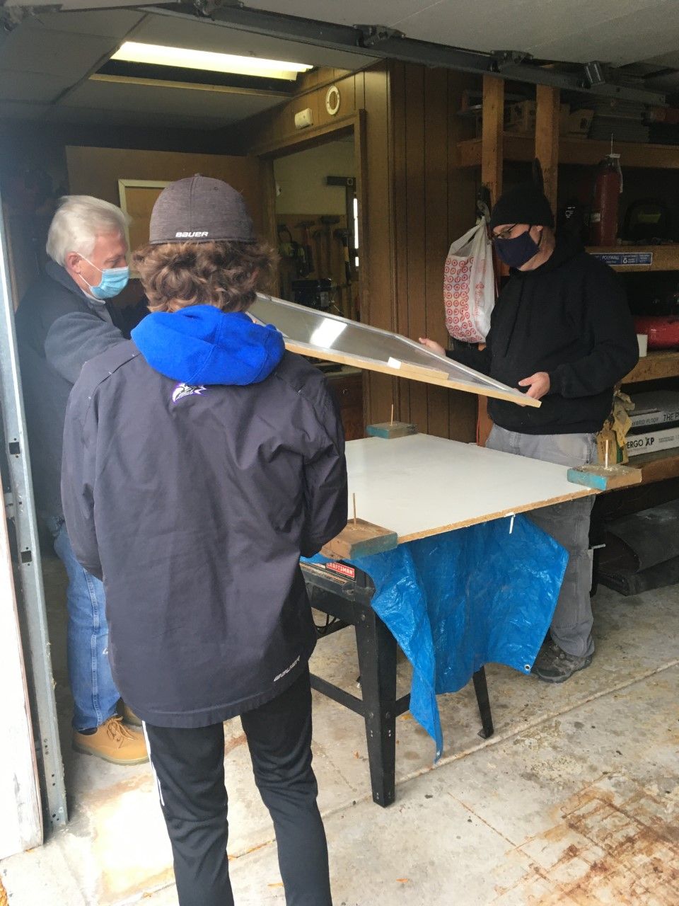 Three people wearing masks examine a framed object inside a garage. One person holds the frame, a second person looks on, and another stands over a table.