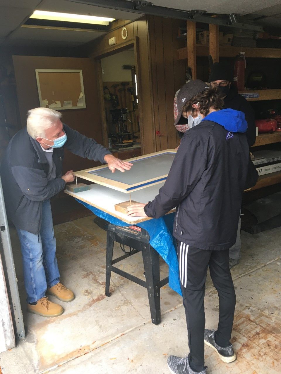 Two people working with a screen, possibly for printmaking, in a workshop. A man in a mask assists a young person.