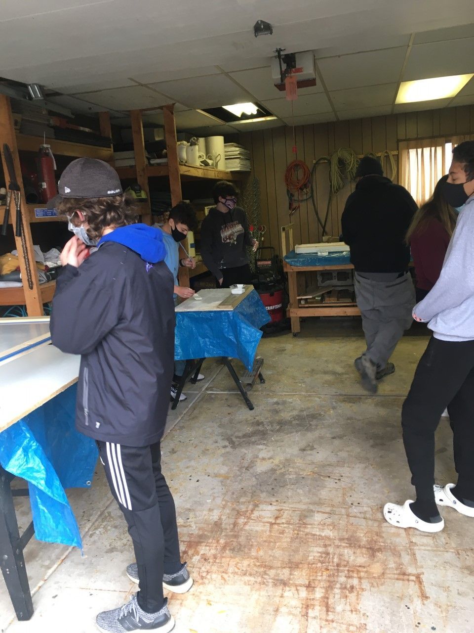 People working in a cluttered garage, some wearing masks, around tables covered with blue tarps.