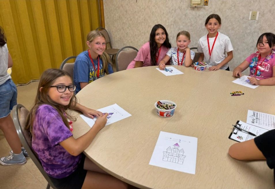 Children seated around a round table, coloring. Some smiling, focused, and holding crayons. Indoors.