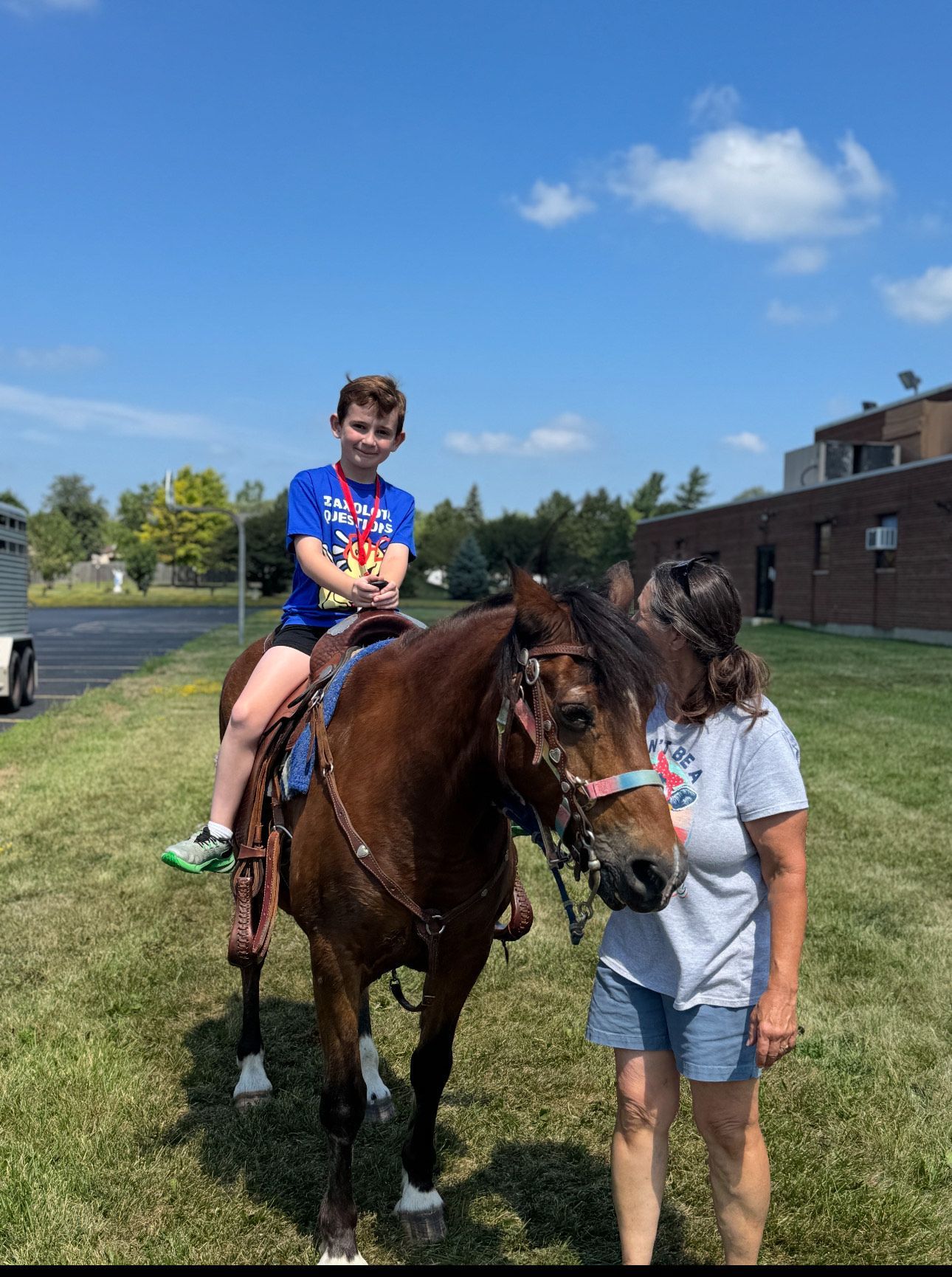 Boy on horseback, brown horse, woman beside, outdoors. Blue sky, green grass.