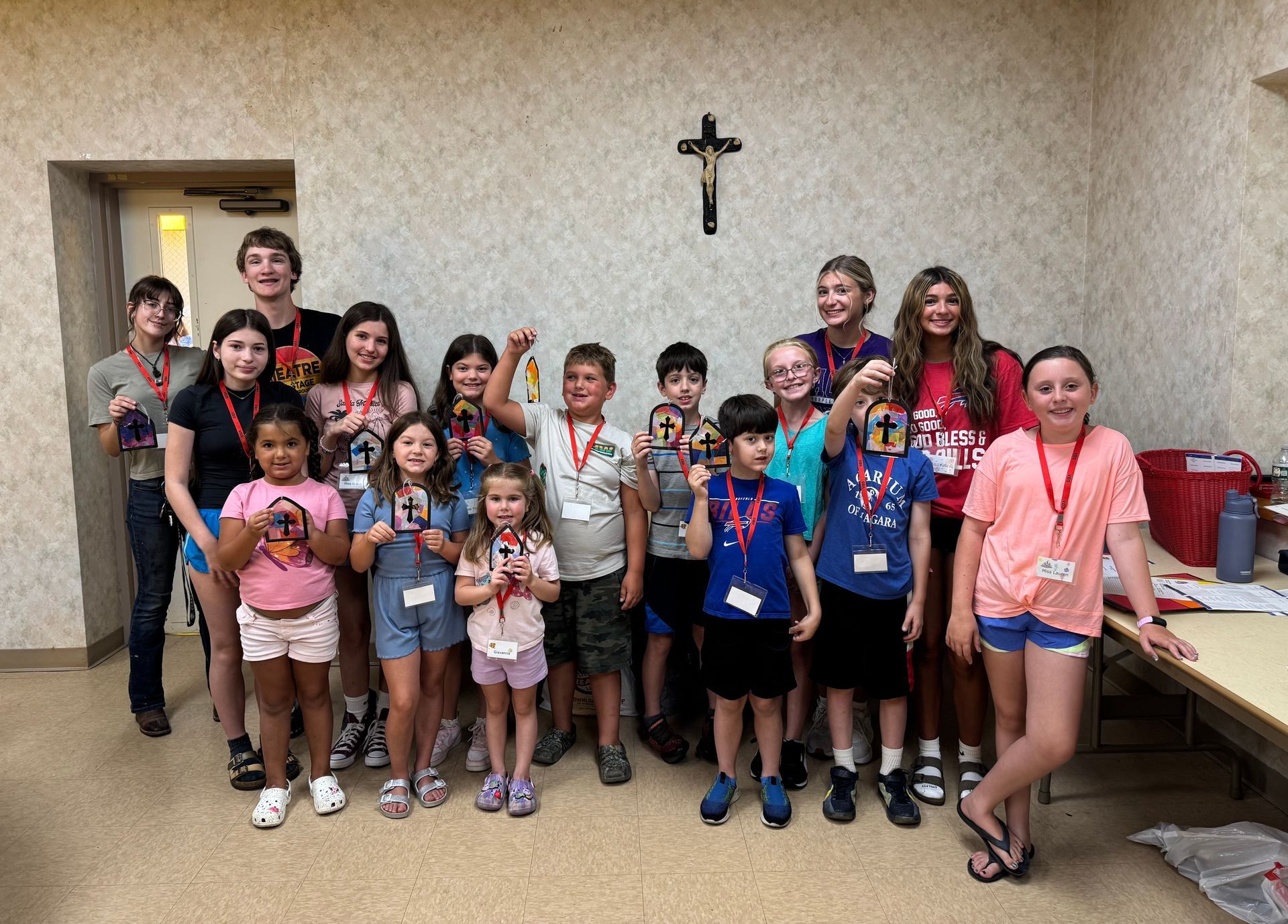 Group of children holding crosses, smiling, in a room with a crucifix on the wall.