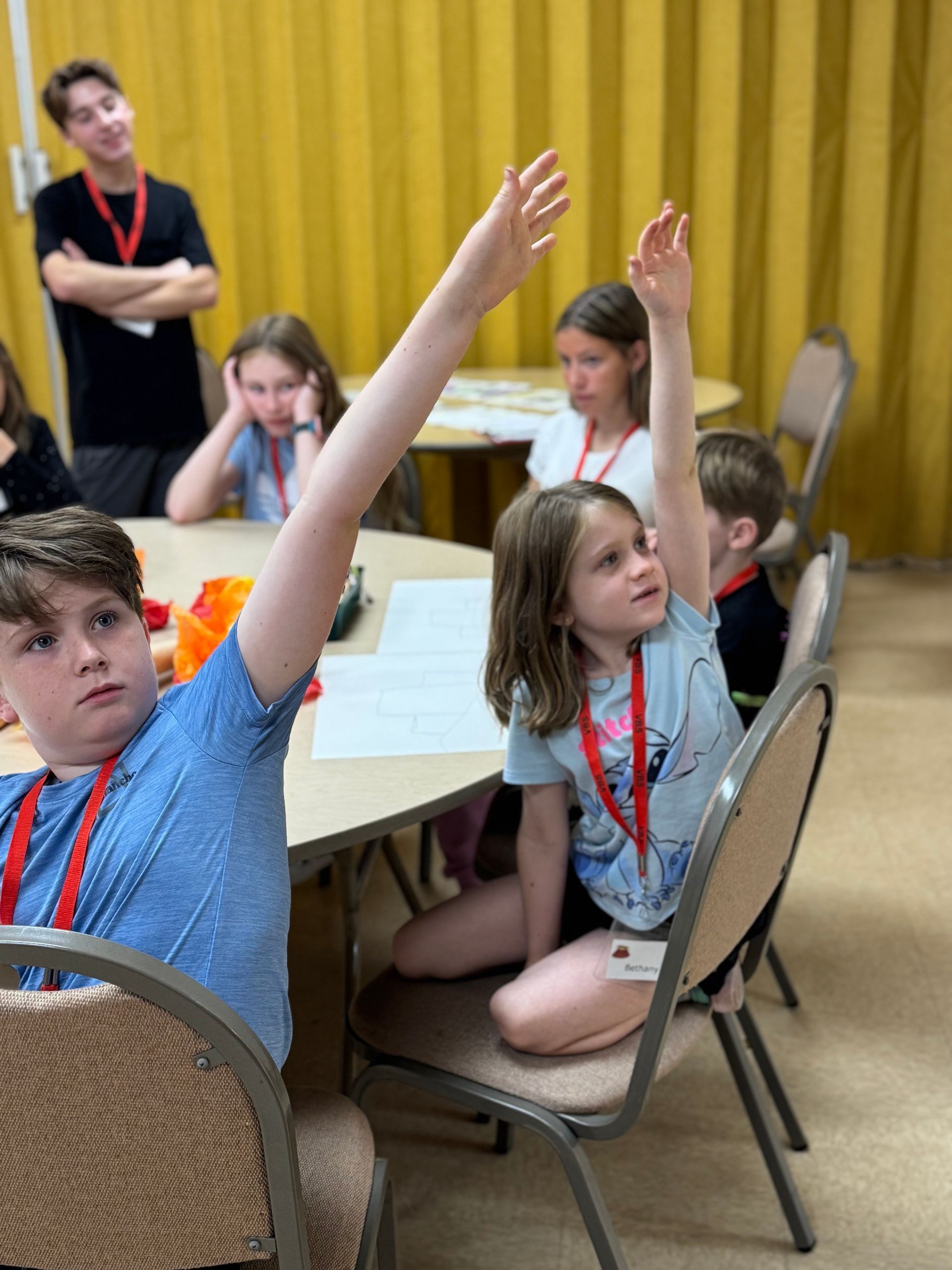 Children raising their hands in a classroom setting. A young adult watches. Yellow curtains in background.