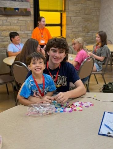 Two boys smiling, making bracelets at a craft table, with other kids and an adult in the background.
