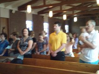 People stand in wooden pews inside a church, some looking forward, others holding babies.