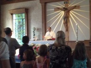 People at a church service. Priest at altar with crucifix. Windows and flowers present.