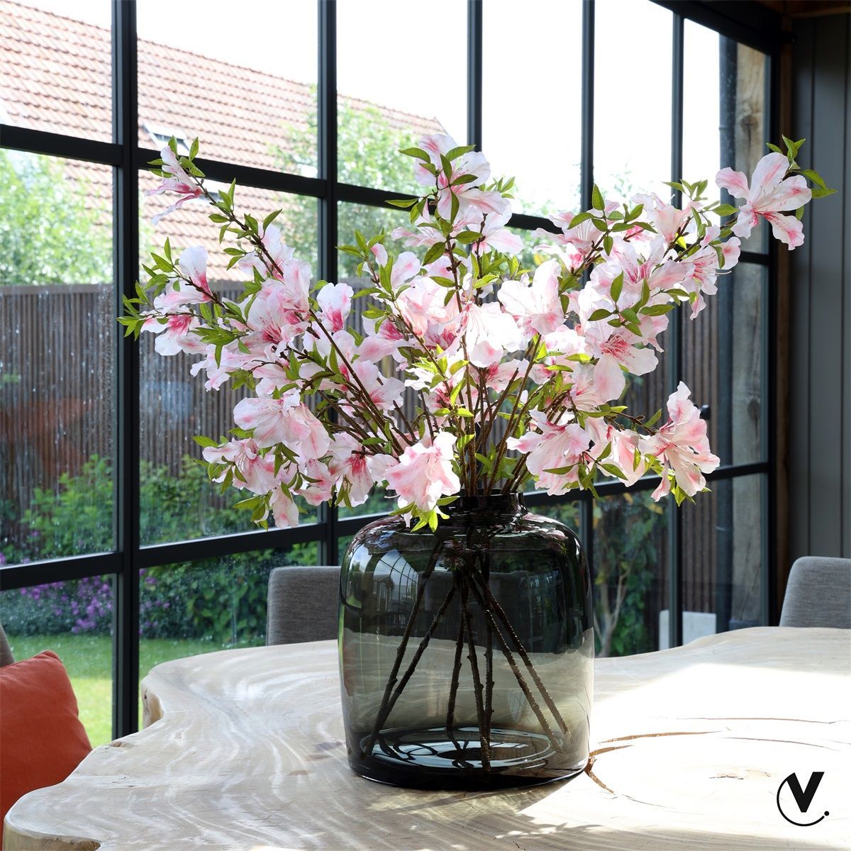 Roze bloesem in een glazen vaas op een tafel bij een zonnig raam.