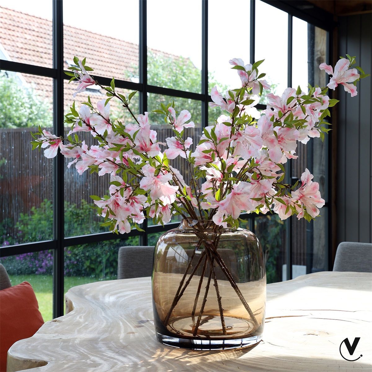 Roze kersenbloesems in een glazen vaas op een eettafel bij een zonnig raam.
