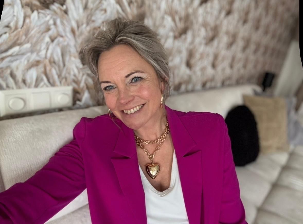 Een lachende vrouw in een magenta blazer zit op een witte bank met een muur met een patroon op de achtergrond.