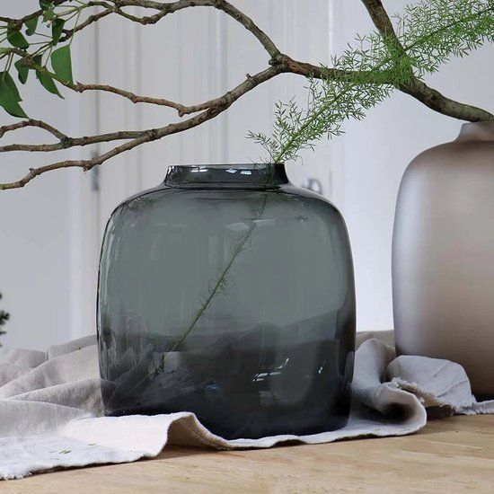 Een rookgrijs glazen vaas bevat groen, op een neutraal linnen tafelkleed, waarbij een tweede vaas gedeeltelijk zichtbaar is.