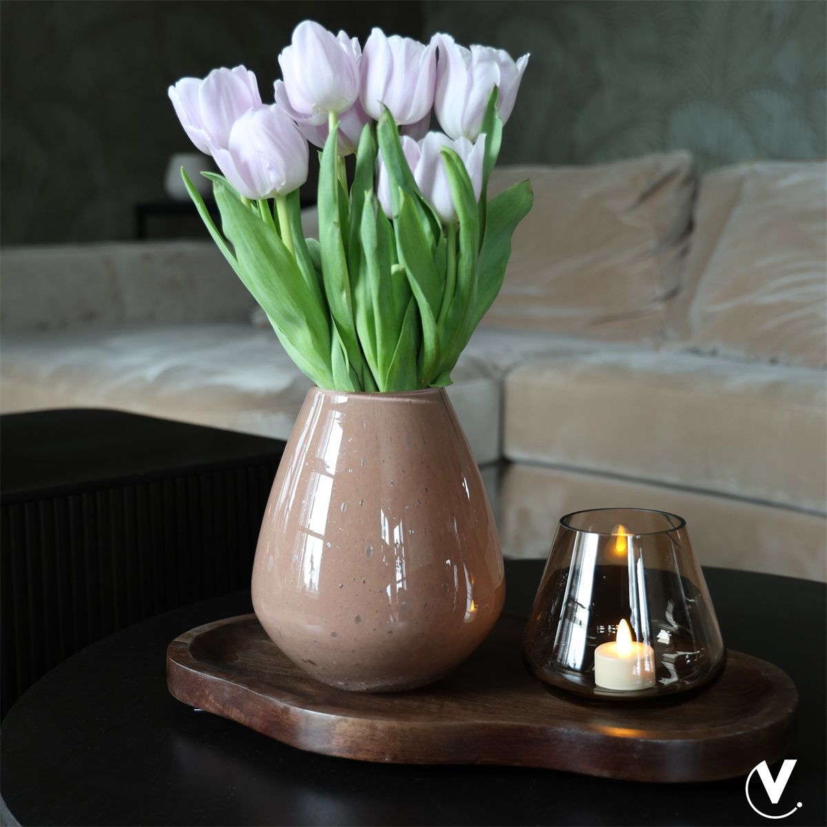 Roze tulpen in een beige vaas op een dienblad naast een brandende kaars op een tafel.