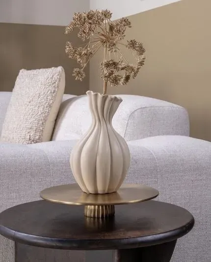 Beige vaas met gedroogde bloemen op een messing schaal op een donkerhouten tafel voor een witte bank.