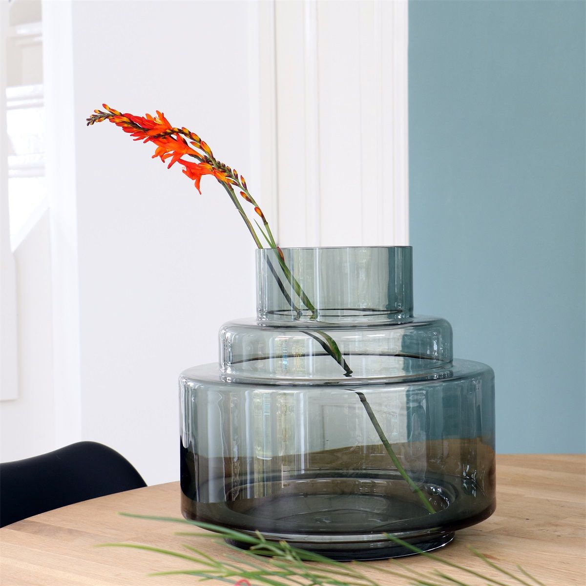 Hoge glazen vaas met oranje bloemen op een houten tafel bij een raam met een blauwe muur.