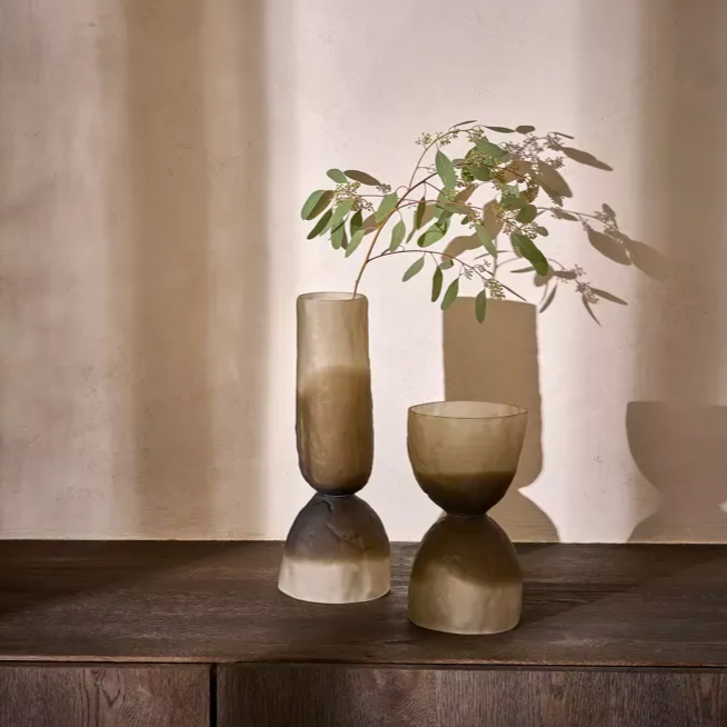 Twee matte glazen windlichten ​​met bladerrijke takken staan ​​op een houten tafel tegen een beige muur.