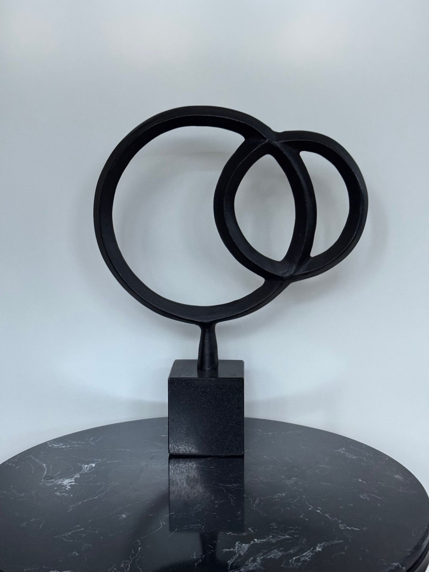 Een sculptuur van zwart metaal, bestaande uit twee elkaar kruisende ringen op een vierkante marmeren sokkel.