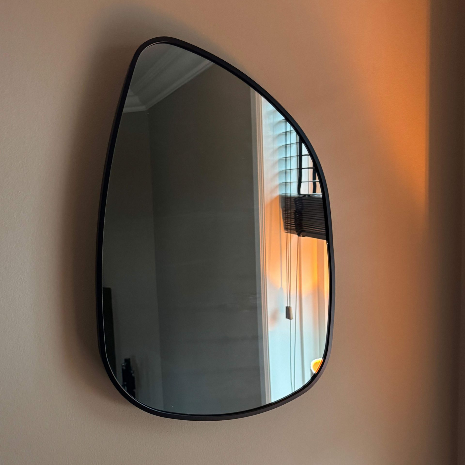 Een zwart omlijste, druppelvormige spiegel aan een beige muur weerspiegelt een zonsondergang en het interieur van de kamer.