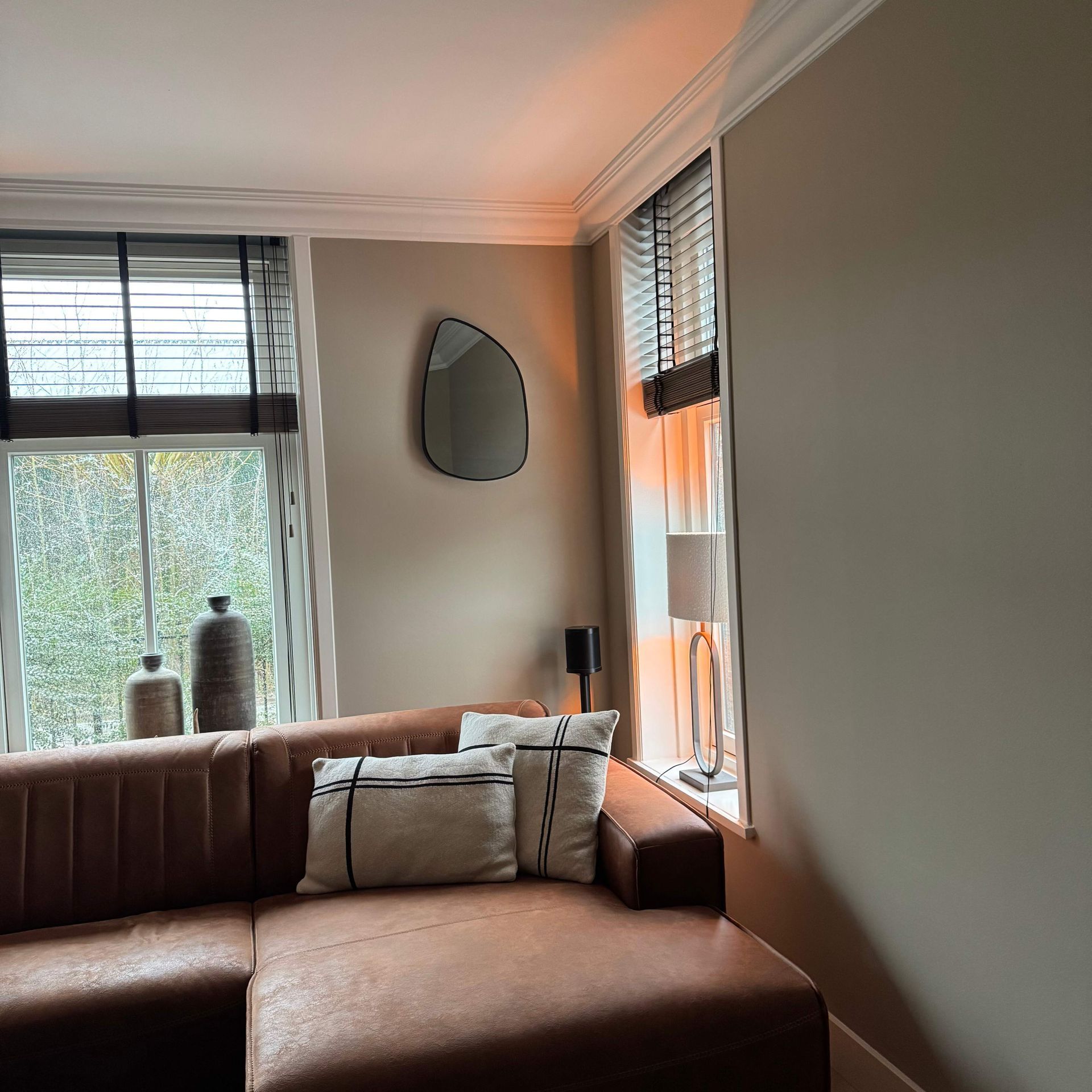 Een zwart omlijste, druppelvormige spiegel aan een beige muur weerspiegelt een zonsondergang en het interieur van de kamer.