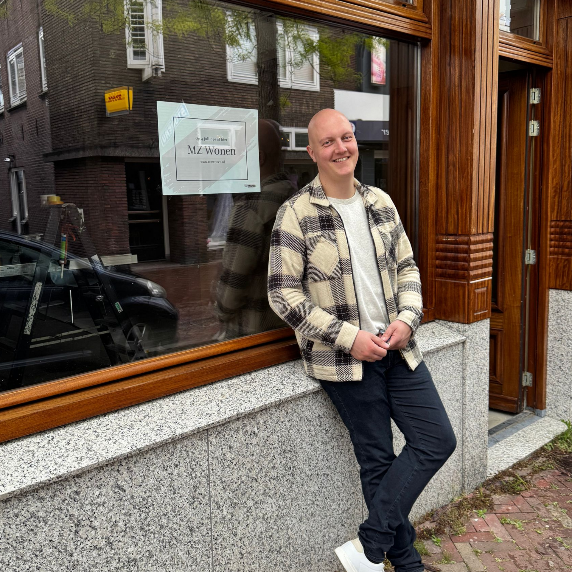 Een persoon leunt tegen een winkelruit, glimlachend, in een zwart-crème gestreepte jas en een donkere broek.